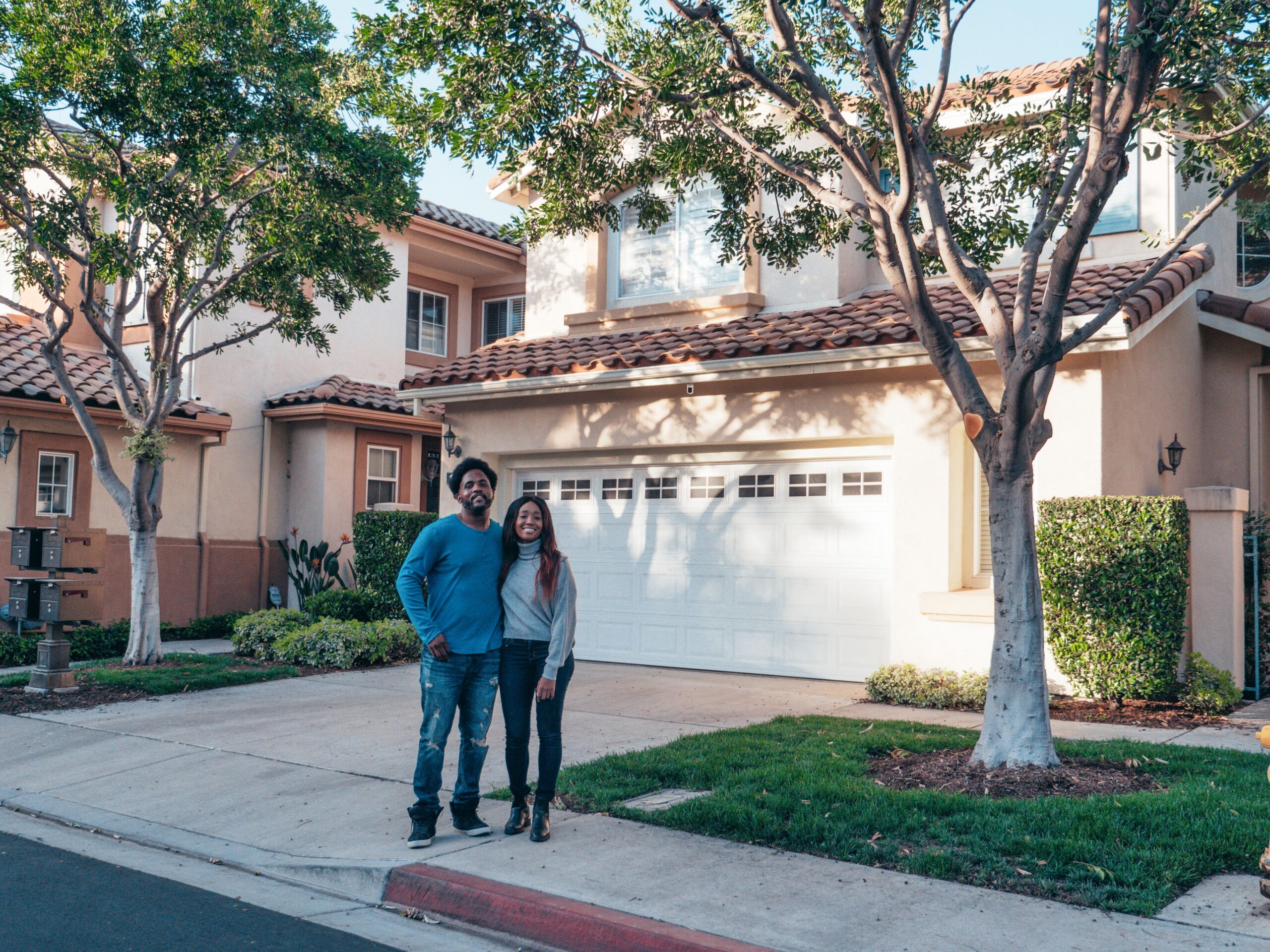 A smiling couple proudly posing in front of their newly purchased house, expressing joy and togetherness.